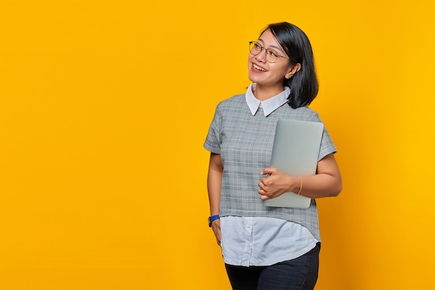Portrait d'une femme asiatique joyeuse portant des lunettes portant un ordinateur portable et regardant de côté sur fond jaune