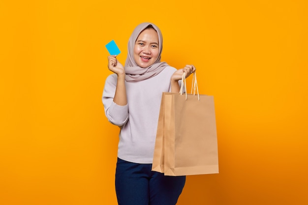 Portrait d'une femme asiatique joyeuse tenant un sac à provisions et montrant une carte de crédit sur fond jaune