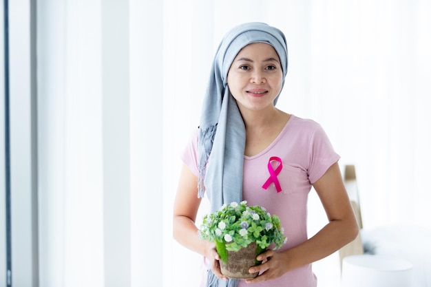 Portrait d'une femme asiatique malade du cancer du sein Tenant des pots de fleurs avec un ruban rose portant un foulard Après un traitement de chimiothérapie à la fenêtre Dans la chambre à coucher à la maison