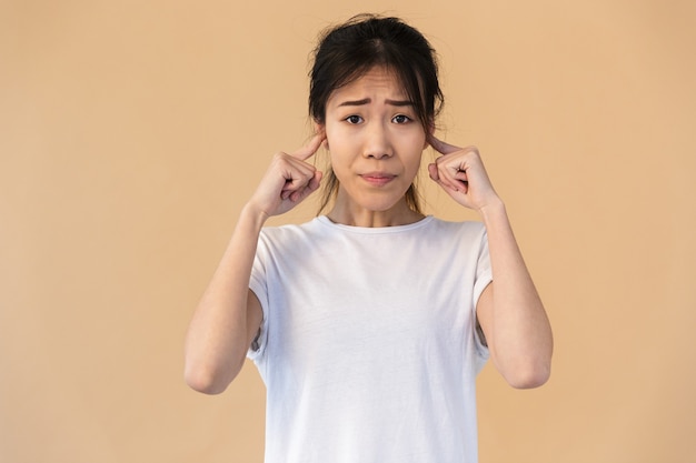 Portrait d'une femme asiatique malheureuse portant un t-shirt basique se bouchant les oreilles avec ses doigts isolés sur un mur beige en studio