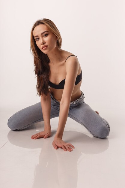 Portrait d'une femme asiatique sexy aux cheveux longs qui pose en lingerie noire et un jean bleu sur fond de studio blanc. tests de modèle de fille maigre en soutien-gorge. jolie femme assise sur le sol sur ses genoux