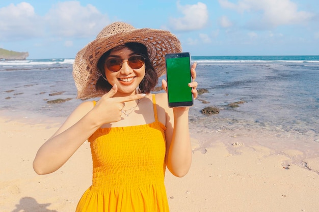 Portrait de femme asiatique souriante heureuse sur la plage montrant l'écran vert pointant du smartphone