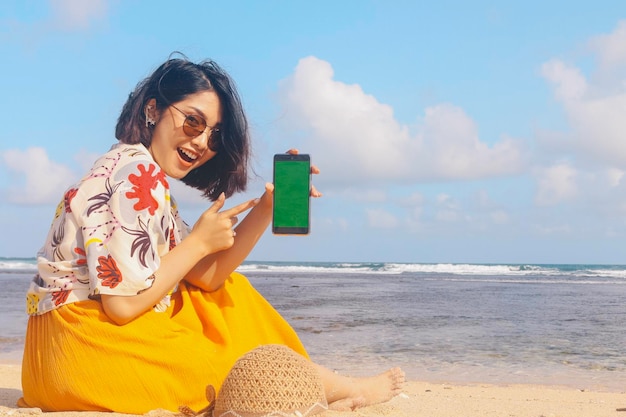 Portrait de femme asiatique souriante heureuse sur la plage montrant l'écran vert pointant du smartphone