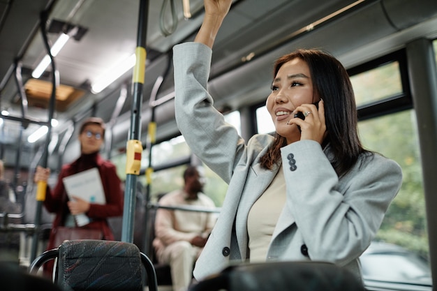 Portrait d'une femme asiatique souriante parlant par smartphone dans un bus et tenant la poignée tout en voyageant...