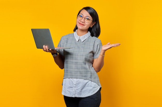 Portrait d'une femme asiatique souriante portant des lunettes tenant un ordinateur portable et montrant un espace de copie dans les paumes sur fond jaune