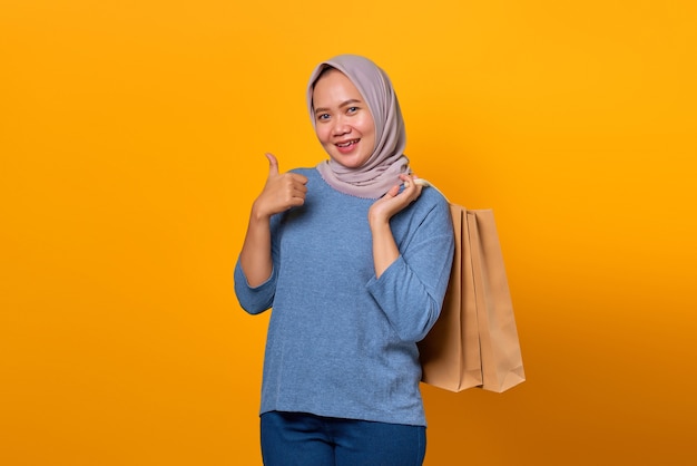 Portrait d'une femme asiatique souriante tenant un sac à provisions et montrant les pouces vers le haut sur fond jaune