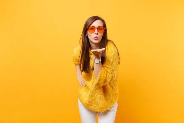 Portrait d'une femme assez tendre en pull de fourrure, lunettes coeur orange soufflant sur les lèvres, envoyant un baiser d'air isolé sur fond jaune vif. Les gens émotions sincères, concept de style de vie. Espace publicitaire.