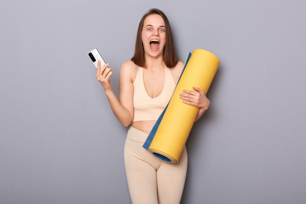 Portrait d'une femme athlétique étonnée et excitée tenant un tapis de fitness enroulé et un téléphone portable hurlant de bonheur en gagnant un abonnement gratuit à la salle de fitness