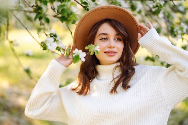 Portrait de femme au chapeau posant près d'un arbre en fleurs Souriante jeune femme appréciant l'odeur des fleurs