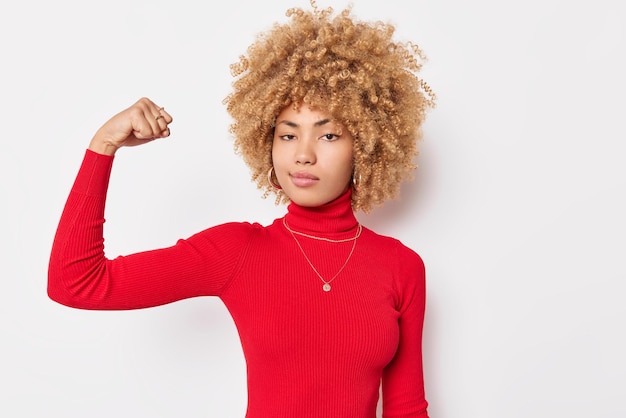 Portrait D'une Femme Aux Cheveux Bouclés Confiante Fléchit Les Biceps Montre Les Muscles Des Bras Regarde Sérieusement La Caméra Porte Des Poses De Col Roulé Rouge Sur Fond Blanc A De L'entraînement Elle Peut Se Défendre