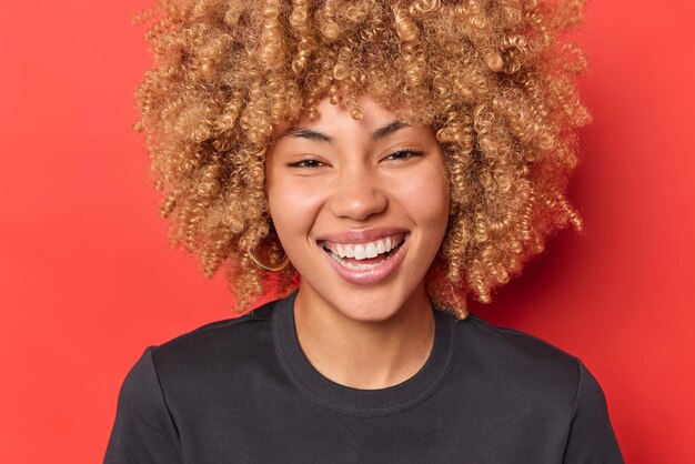 Portrait d'une femme aux cheveux bouclés positive sourit sur la route montre des dents blanches étant de bonne humeur entend quelque chose de drôle habillé avec désinvolture isolé sur fond rouge vif Concept d'émotions heureuses