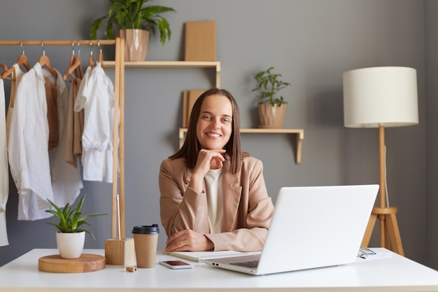 Portrait d'une femme aux cheveux bruns souriante et satisfaite portant une veste beige assise sur son lieu de travail devant un ordinateur portable regardant la caméra tenant le menton exprimant des émotions positives sur son travail
