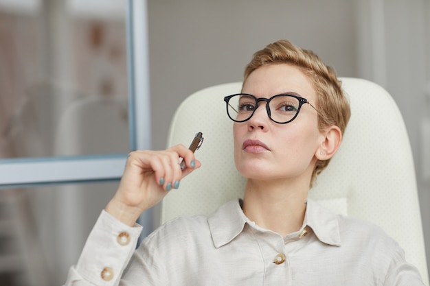 Portrait de femme aux cheveux courts moderne portant des lunettes à la recherche de suite pensivement tout en se prélassant dans une chaise de bureau au travail, copiez l'espace