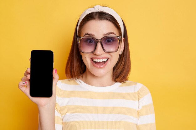 Portrait d'une femme aux cheveux noirs excitée surprise portant une chemise rayée posant isolé sur fond jaune montrant un téléphone portable avec écran vide avec espace de copie pour la publicité