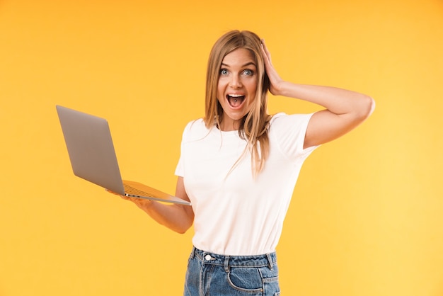 Portrait d'une femme blonde étonnée portant un t-shirt décontracté exprimant son émerveillement tout en utilisant un ordinateur portable isolé sur un mur jaune en studio