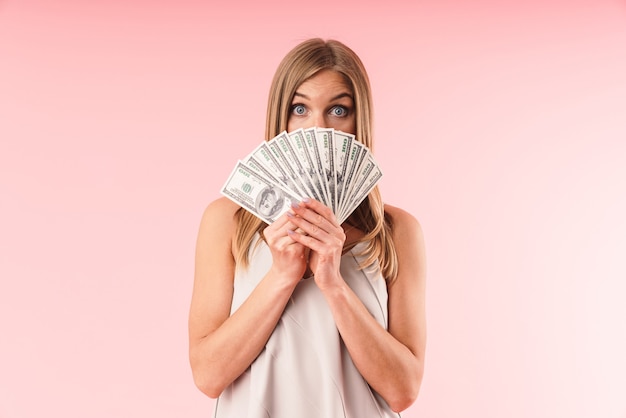 Portrait d'une femme blonde excitée regardant à l'avant tout en se couvrant le visage avec des billets d'argent isolés sur un mur rose en studio