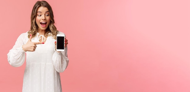 Portrait d'une femme blonde heureuse excitée et étonnée en robe blanche pointant le doigt sur l'écran du téléphone portable regardant le smartphone avec un sourire heureux et surpris sur fond rose