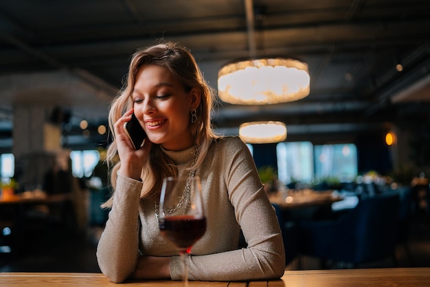 Portrait d'une femme blonde joyeuse parlant au téléphone portable assise à table avec du vin rouge dans un restaurant moderne à l'intérieur sombre