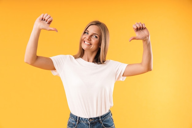 Portrait d'une femme blonde joyeuse portant un t-shirt décontracté souriant et pointant du doigt elle-même isolée sur un mur jaune en studio