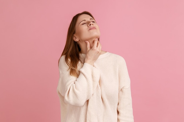 Portrait d'une femme blonde malade touchant un mal de cou se sentant mal souffrant d'inflammation des amygdales concept médical portant un chandail blanc Prise de vue en studio intérieure isolée sur fond rose