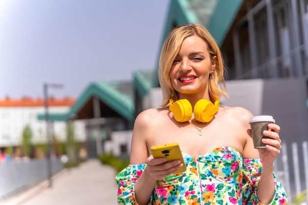 Portrait d'une femme blonde marchant avec le téléphone à travers la ville avec des écouteurs jaunes et un style de vie de café à emporter d'un modèle dans la ville