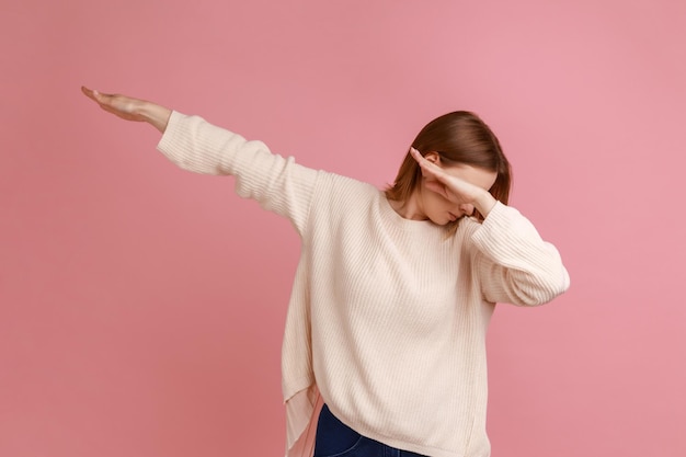 Portrait d'une femme blonde montrant une pose de danse dab, célèbre mème Internet de triomphe, exécutant des tendances de dabbing avec un geste de la main, portant un pull blanc. Tourné en studio intérieur isolé sur fond rose.