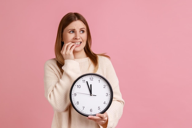 Portrait d'une femme blonde nerveuse tenant une grande horloge murale et se mordant les ongles, a besoin de plus de temps pour le travail, date limite, portant un pull blanc. Tourné en studio intérieur isolé sur fond rose.