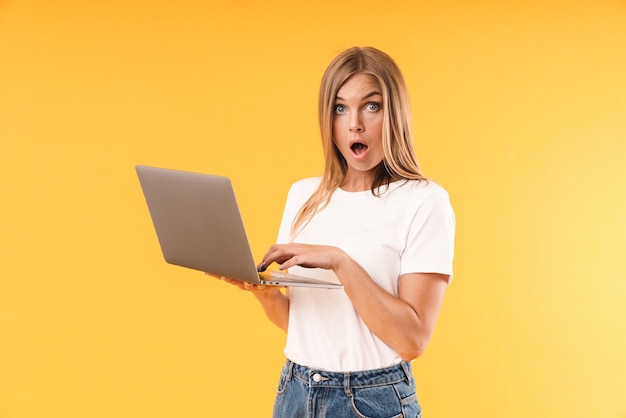 Portrait d'une femme blonde perplexe portant un t-shirt décontracté exprimant son émerveillement tout en utilisant un ordinateur portable isolé sur un mur jaune en studio