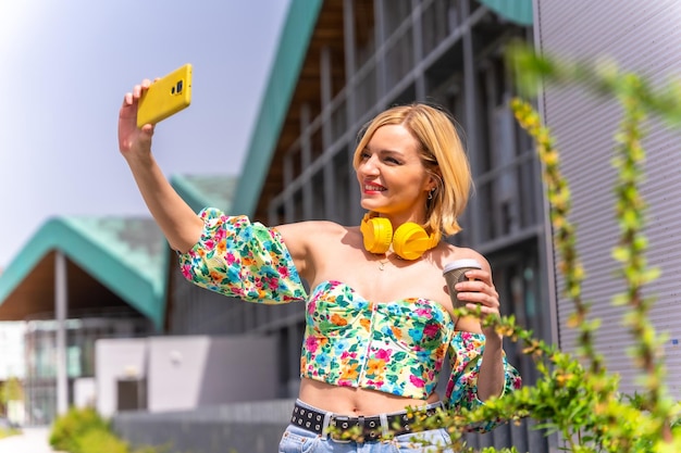 Portrait d'une femme blonde prenant un selfie avec son téléphone dans la ville avec des écouteurs jaunes et un style de vie de café à emporter d'un modèle dans la ville