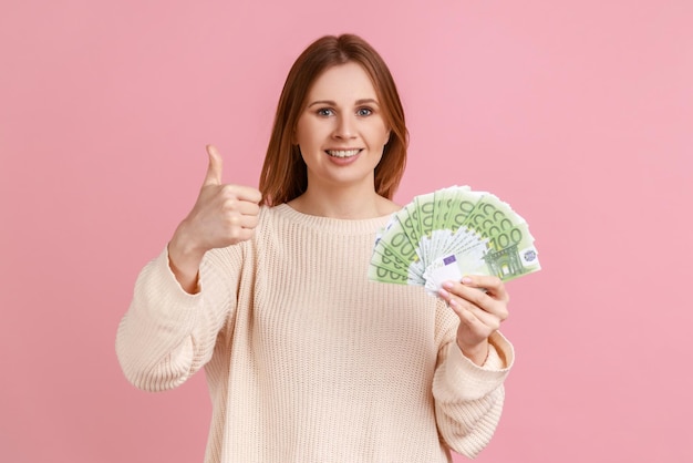 Portrait d'une femme blonde souriante positive tenant de nombreux billets en euros et montrant le pouce vers la caméra, satisfaite de gagner à la loterie, portant un pull blanc. Studio intérieur tourné isolé sur fond rose