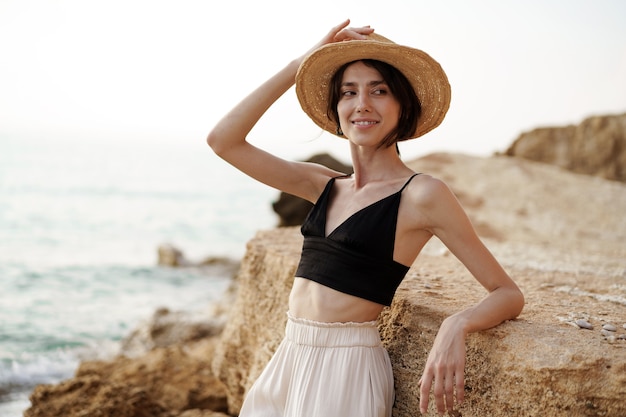 Portrait de femme en bralette noire et pantalon blanc s'appuyant sur un rocher à la plage