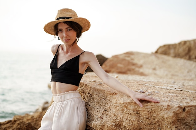 Portrait de femme en bralette noire et pantalon blanc s'appuyant sur un rocher à la plage