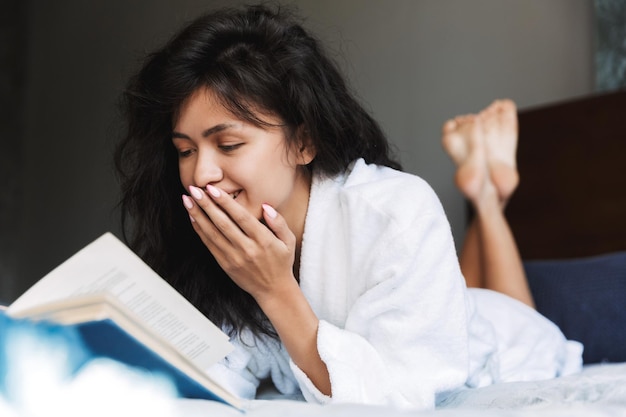 Portrait d'une femme brune excitée portant un livre de lecture de peignoir au lit à la maison