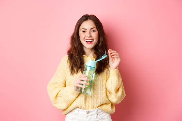 Portrait d'une femme brune heureuse avec une coiffure frisée, de l'eau potable avec du citron de bouteille personnelle, riant et souriant, se sentant en bonne santé, debout contre le mur rose.