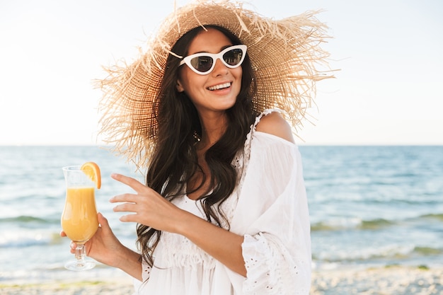 Portrait d'une femme brune heureuse dans des lunettes de soleil et un chapeau de paille souriant en marchant au bord de la mer avec un verre de jus d'orange