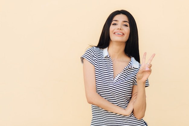 Portrait de femme brune joyeuse montre un geste de paix, maintient deux doigts levés