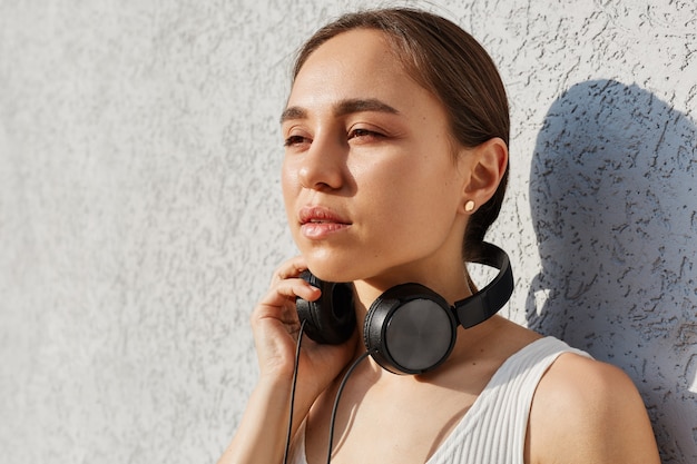 Portrait d'une femme brune rêveuse vêtue d'un haut blanc et ayant des écouteurs sur le cou, détournant les yeux avec une expression faciale pensive, posant contre un mur gris en plein air.