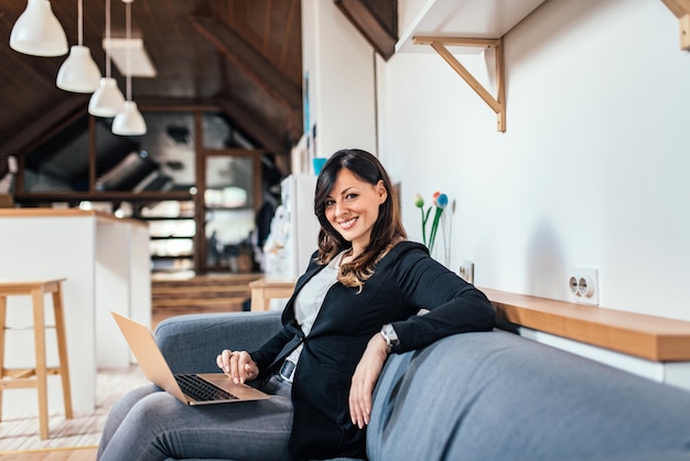 Portrait d&#39;une femme brune souriante à l&#39;aide d&#39;un ordinateur portable assis sur le canapé. Regardant la caméra.