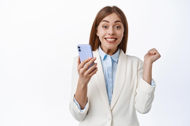 Portrait d'une femme de bureau chanceuse en costume d'affaires sautant du triomphe et de l'excitation tenant un smartphone se réjouissant et disant oui gagnant en ligne fond blanc