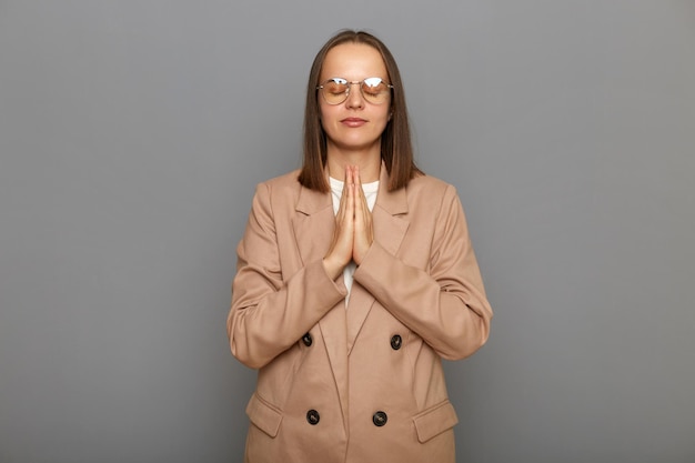 Portrait d'une femme calme et détendue aux cheveux noirs portant une veste debout dans les mains de la paume et faisant de la méditation de yoga les yeux fermés contre un mur gris essayant de se détendre pendant son travail