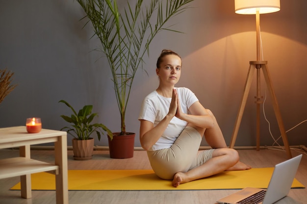 Portrait d'une femme calme et détendue avec un chignon en t-shirt pratiquant le yoga à l'aide d'un ordinateur portable pour travailler avec son canapé en ligne assis sur le sol, main dans la main en geste de namaste et méditant relaxant