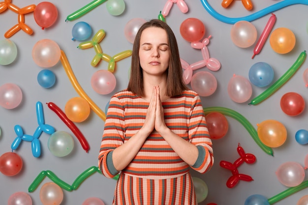 Photo portrait d'une femme calme reconnaissante aux cheveux bruns portant une robe rayée posant sur fond gris avec des ballons gonflés se relaxant avec du yoga priant avant le dîner de fête