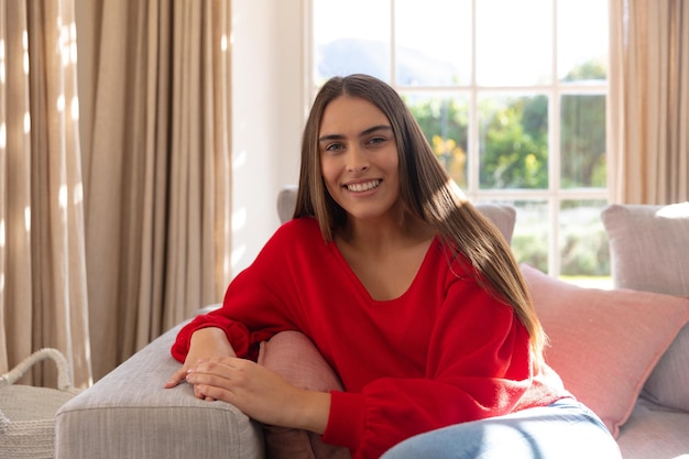 Portrait d'une femme caucasienne heureuse assise sur un canapé dans un salon ensoleillé, souriant à la caméra. Concept de vie domestique, de loisirs et de bonheur.