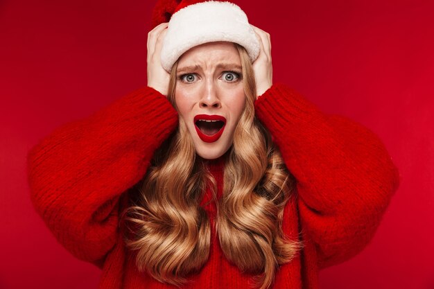 Portrait d'une femme caucasienne hurlante posant isolée sur un mur rouge avec un rouge à lèvres rouge vif vêtu d'un pull rouge chaud portant un chapeau de noël.