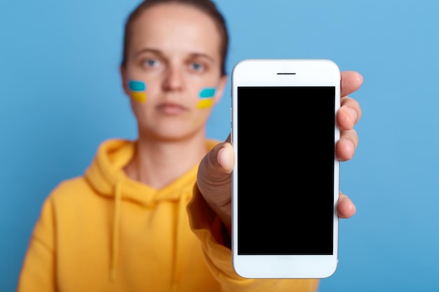 Photo portrait d'une femme caucasienne portant un sweat à capuche jaune et avec le drapeau de l'ukraine sur sa joue montrant un téléphone portable avec un écran vide avec un espace pour la publicité isolé sur fond bleu