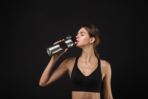 Portrait de femme caucasienne de remise en forme dans l'eau potable de vêtements de sport de la bouteille pendant l'entraînement dans la salle de sport isolée sur fond noir