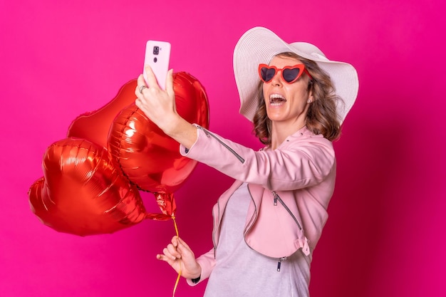 Portrait d'une femme caucasienne s'amusant avec un chapeau blanc dans une boîte de nuit avec des ballons coeur prenant un fond rose selfie