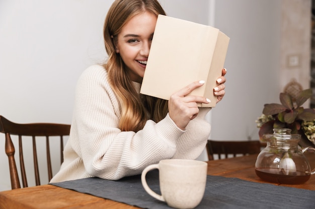 Portrait d'une femme caucasienne souriante portant des vêtements décontractés, lisant un livre et buvant du thé dans une chambre confortable