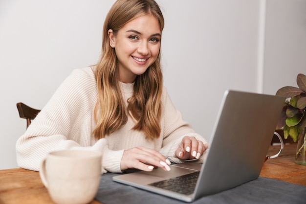 Portrait d'une femme caucasienne souriante portant des vêtements décontractés utilisant un ordinateur portable et buvant du thé dans une chambre confortable