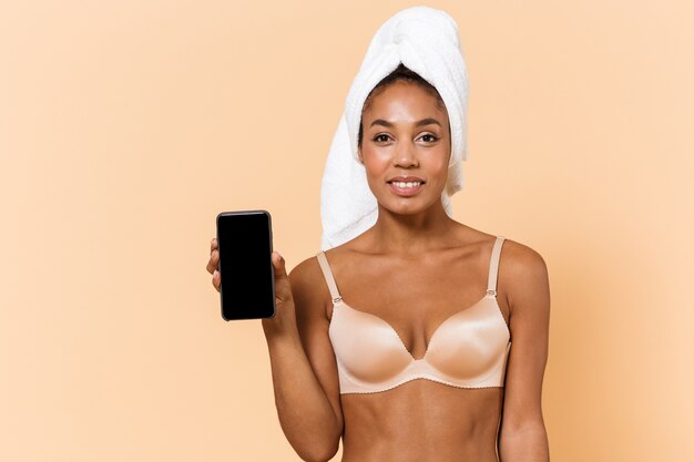 Portrait de femme charmante enveloppée dans une serviette et portant de la lingerie blanche à l'aide de téléphone mobile, isolé sur mur beige